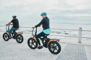 Couple, helmet or electrical bike by sea or ocean beach in bonding transport, clean energy or sustainability travel. Technology, electric or eco friendly bicycle for happy woman or mature cycling man.